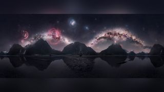 Person stands on a rocky shore before reflective water, jagged mountains, and a vivid star-filled galaxy sky