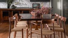 Image of a wooden dining table with 10 wooden dining chairs around it. There is a beige/brown stripped rug under the table and a bowl of pink flowers on top.