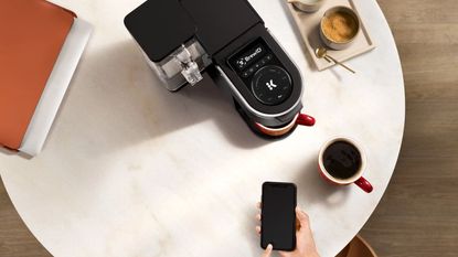 An aerial view of the he Keurig K-Supreme SMART Coffee Maker being controlled by a smartphone next to a cup of coffee