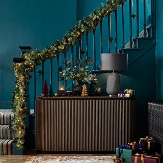 Hallway and staircase dressed for Christmas with lit garland running up bannisters and Christmas tree miniatures on dark wood sideboard