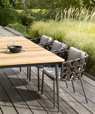 A gray wooden deck with table and chairs, bordered by long grasses.