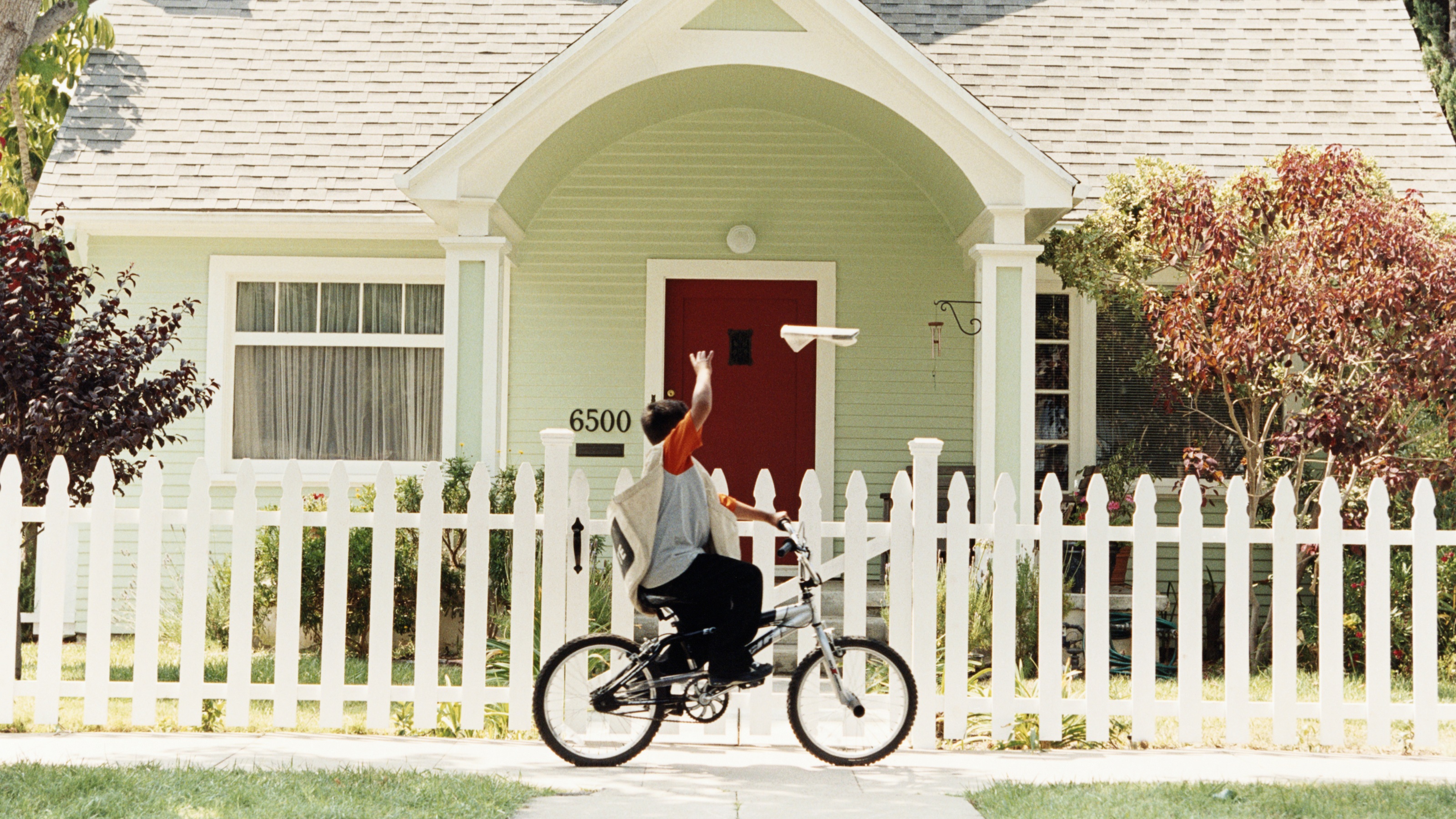 A paper boy on a bike throws a newspaper toward the front porch of a house.