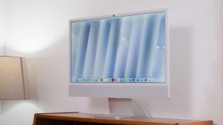 Apple iMac on a wooden desk against a white wall next to a lamp