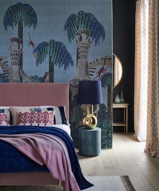 A luxurious blue bedroom with a blue mural of a jungle at twilight, pink bed, dark blue and pink soft furnishings and a gold bedside lamp.