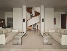 A spiral staircase fitted centrally between two pillars in the living space, with two long glass coffee tables and two cream sofas.