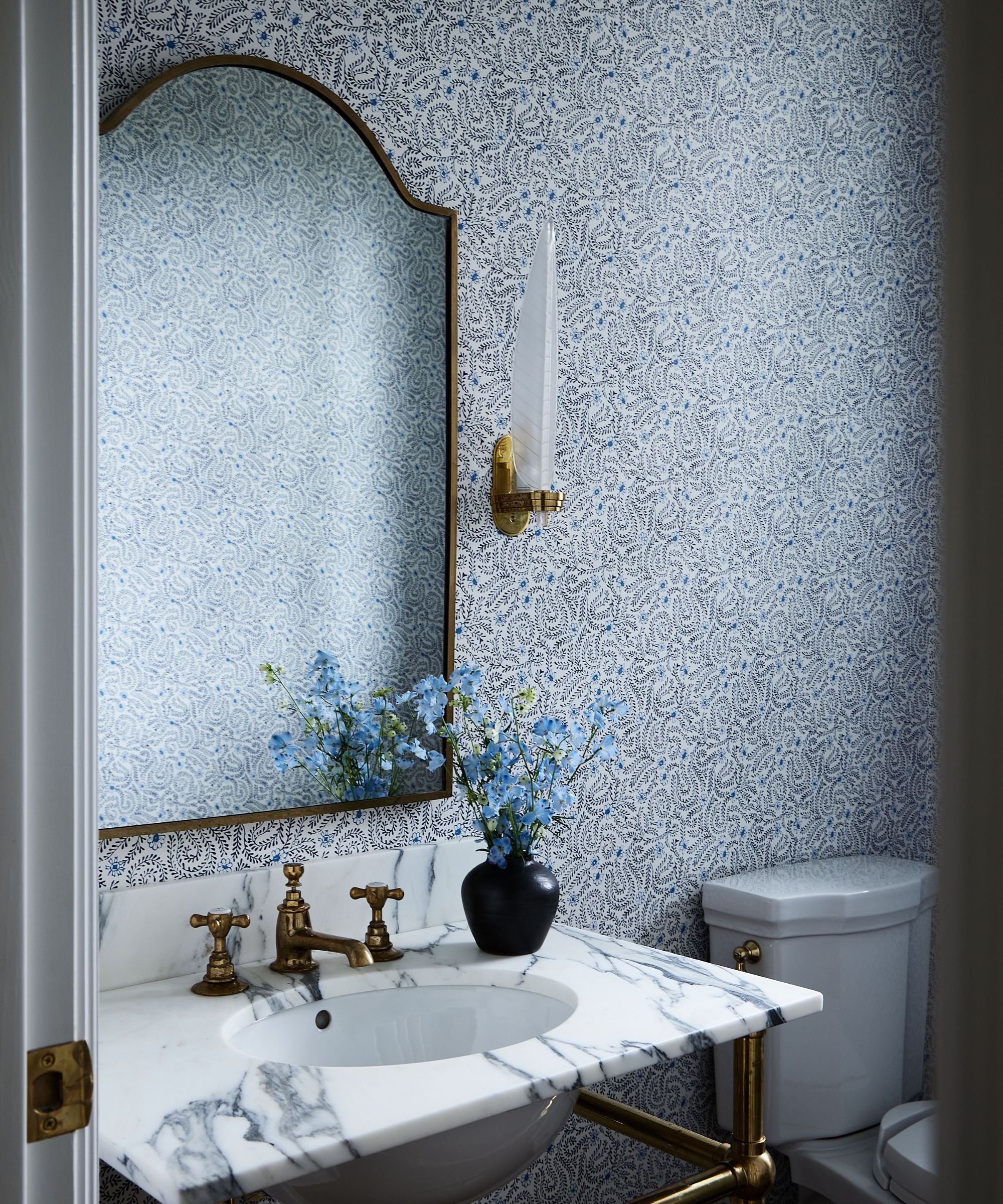 Blue and white bathroom with ditsy wallpaper, marble sink, curved mirror, gold sconces and vase with blue flowers