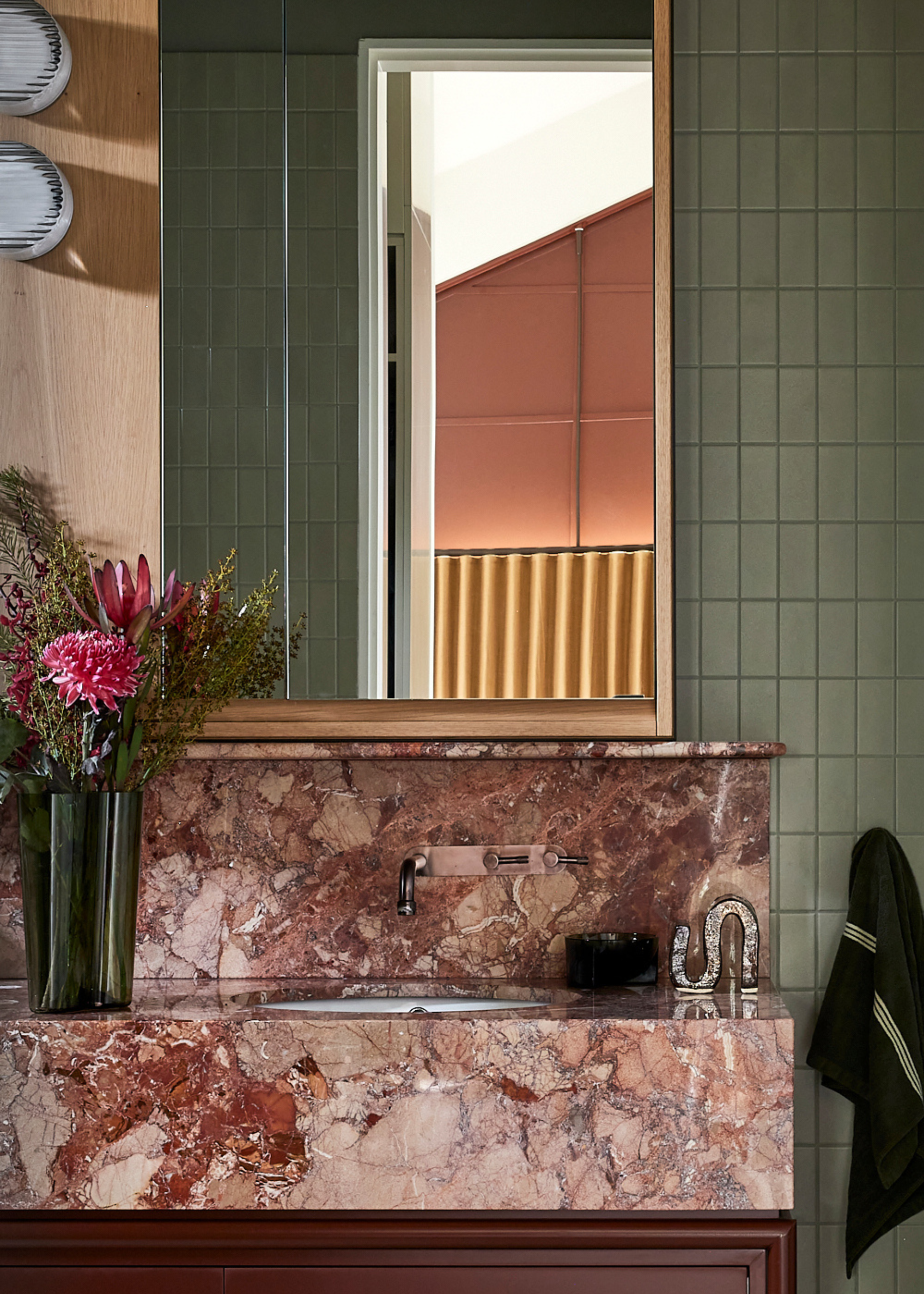 A bathroom with green tiles, a gold framed mirror, a pink stone sink, a green vase of flowers, and silver hardware