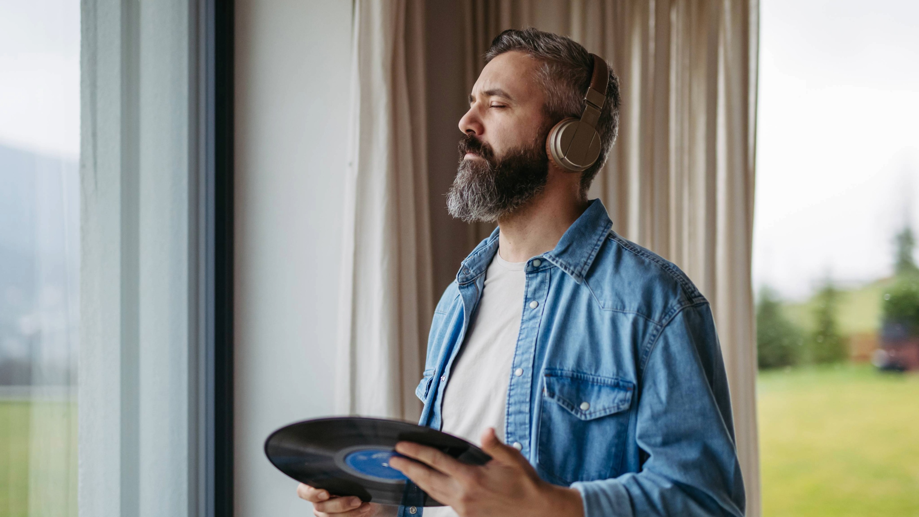 Handsome man listening to vinyl with headphones, standing by window