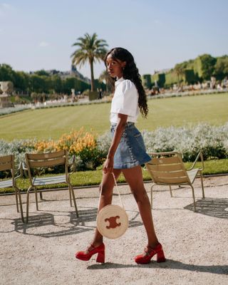 Influencer wearing white shirt, denim mini skirt, and red platform Mary Janes while walking in garden.