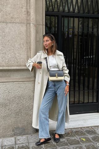 spring trench coats shown in a photo of a French woman standing outside leaning against a wall wearing black sunglasses, gold hoops, a trench coat, a white tank top, a two-tone crossbody shoulder bag, straight-leg jeans, and black ballet flats