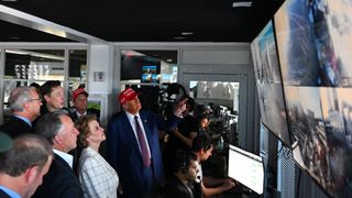 a crowd of well-dressed people stand in a mission control room while engineers stare at their computer screens