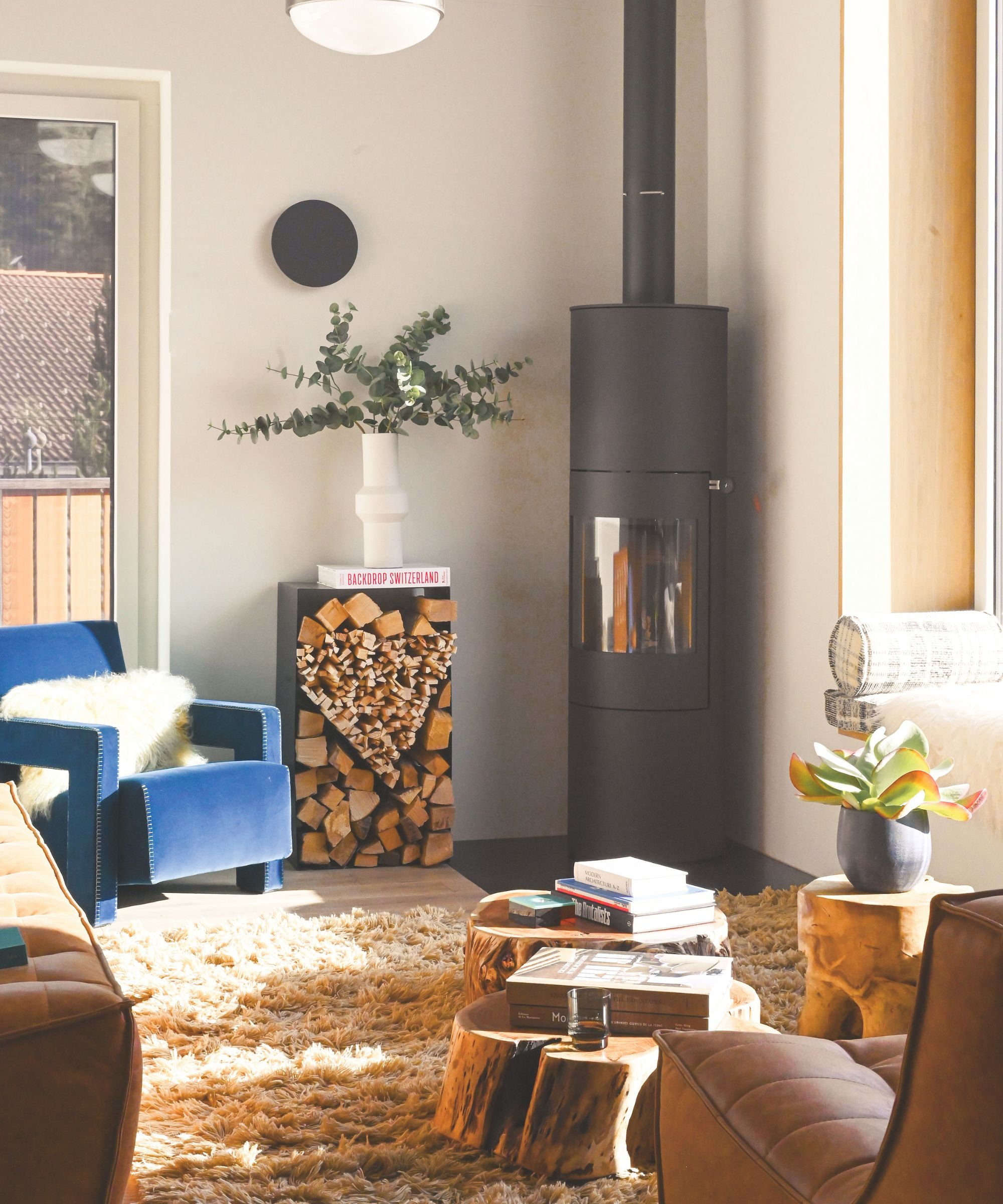 A cozy living room corner with a black cylindrical wood-burning stove, a stack of firewood, a blue velvet armchair, and natural wood stump coffee tables on a shag rug