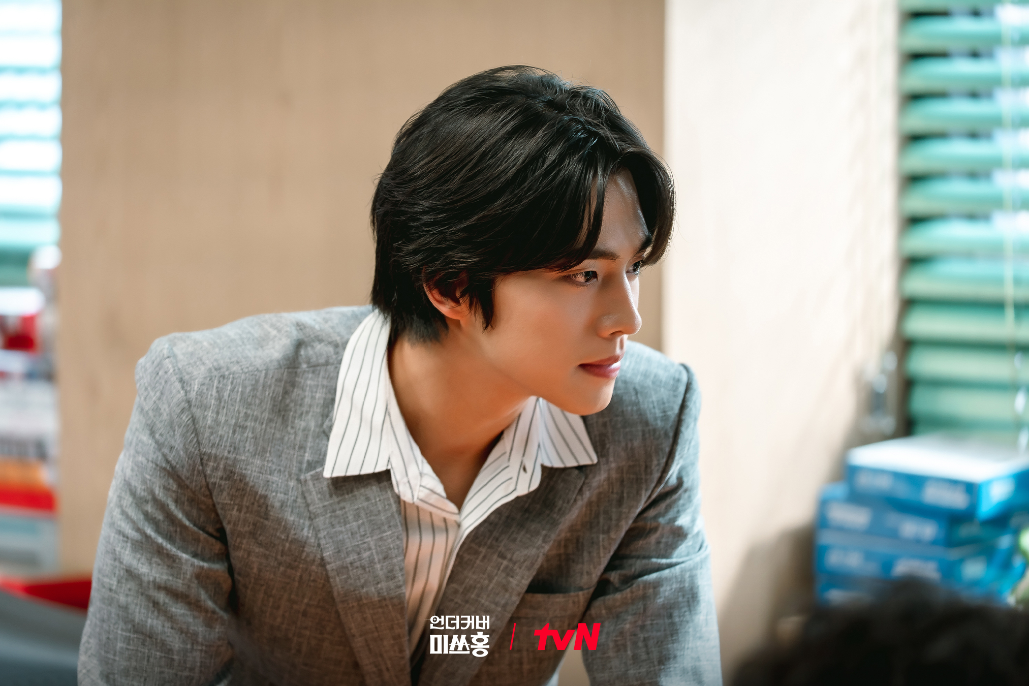 A Korean man wearing a gray suit (Cho Han-gyeol as Albert Oh) leans on a desk in an office, in the Netflix K-drama 'Undercover Miss Hong.'