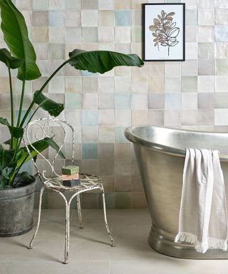 A bathroom with light gray tiled walls with a black and white wall art print, a tall dark green plant, a silver chair, and a large silver bathtub