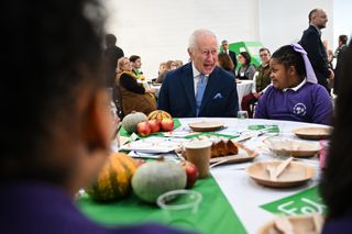 King Charles sitting at a table with school children wearing purple uniforms while talking and laughing