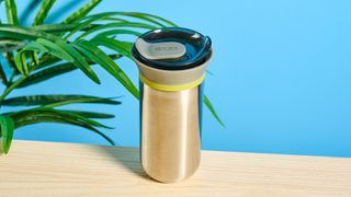 Wacaco Cuppamoka, a stainless steel coffee pour-over brewer, photographed against a blue background, with brown filters and a sippy lid