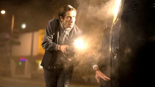Jake Gyllenhaal as Louis Bloom holding a flashlight and crouching next to a car at night in the thriller movie Nightcrawler.