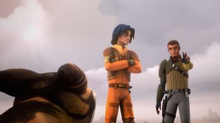Screenshot from the animated T.V. show Star Wars Rebels. Here we see Ezra Bridger as a teenager. HE has shoulder-length dark blue hair and he is wearing an orange jumpsuit with brown shoulder pads and belt. His arms are crossed as he's looking over at the adult to his right, who is Jedi Kanan Jarrus. Kanan has short brown hair, a goatee and is wearing gray trousers, beige shirt and an olive-green protective guard fitted along his whole right arm. Kanan's hand is outstretched palm facing forwards pointing towards a small dog-sized critter in front of them.