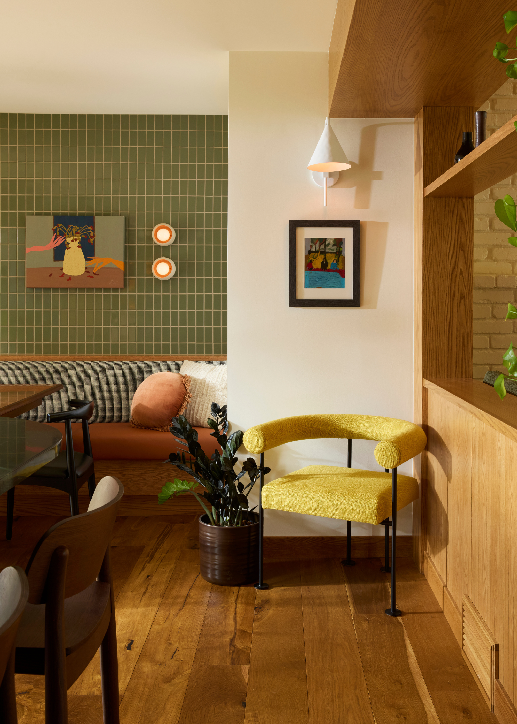A living room corner with banquette seating a tiled green wall, a pair of wall sconces and a painting beside a white wall with a yellow armchair, a white wall sconce and a potted jade plant in a brown planter