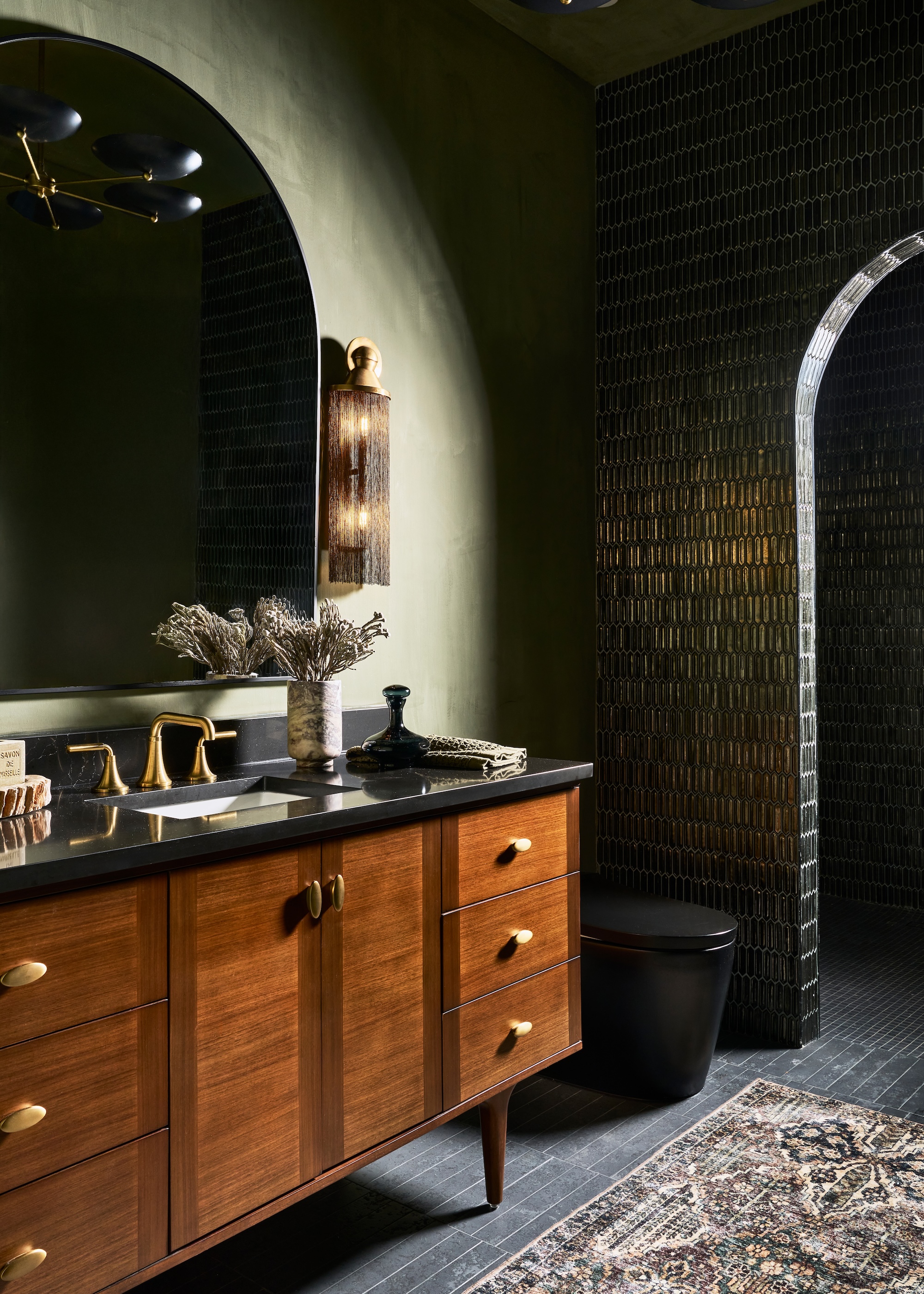 a dark and moody green and brass bathroom shown with a vintage style sideboard vanity basin and oversized curved mirror