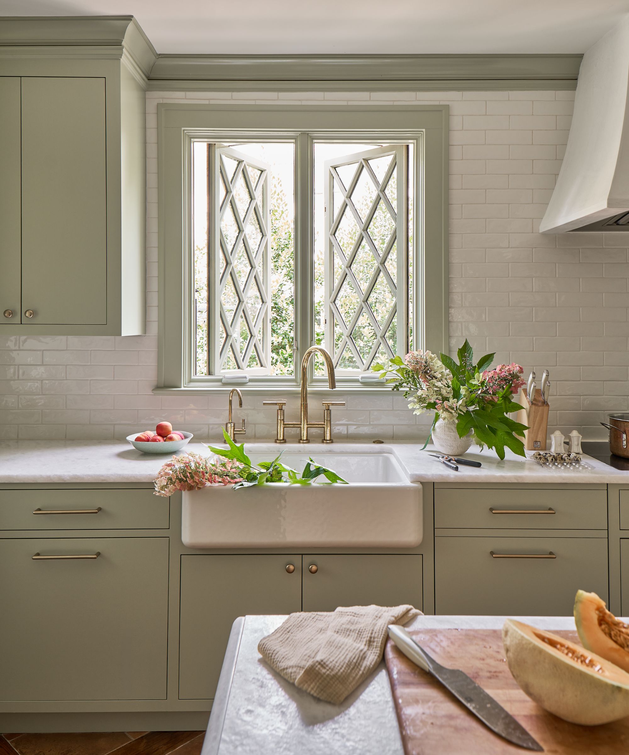 A modern rustic kitchen with pale green cabinets, white walls talls and countertop, a Belfast sink with brass taps in front of a Tudor window, and pink flowers on the side and in the sink.