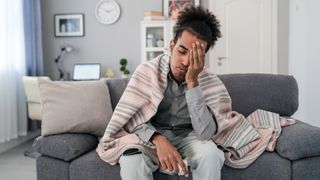 Young african american man, sitting on the sofa at home, having a fever and headache and touching his forehead to check his temperature.