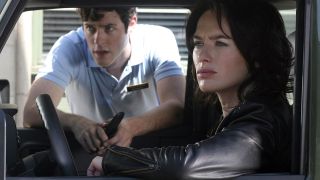 Still from a Terminator movie. A woman with dark shoulder-length hair wearing a black leather jacket is sitting behind the wheel of a large car. Leaning in the window is a man with short dark hair and he is wearing a light blue polo shirt with a nametag. They both are staring into the distance with a look of concern on their faces.