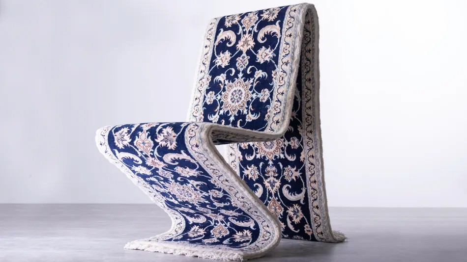 A single chair made from a large, ornate, blue-and-white patterned rug is positioned against a clean white and gray background.