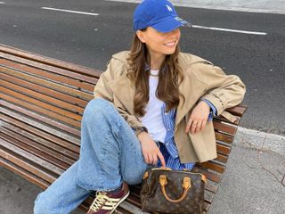 Fashion influencer Luna Isabella wearing a chic spring outfit styled with on-trend basics, including faded blue jeans, a white tee, a striped button-down shirt, a cropped trench, a baseball cap, a top-handle bowling bag, and vintage-inspired Adidas SL 72 OG sneakers.