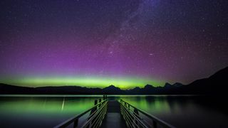 a lake reflects the green hue of the aurora borealis, or northern lights, shining from the sky above. In the sky there are purple bands of light as well as green and the milky way stretching up through the center of the image.