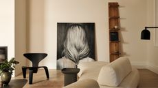 modern artwork with large artwork leaning against the wall, black chair, beige sofa in the middle of the room, black side table, timber bookshelf leaning against wall, and coffee table with vase of foliage