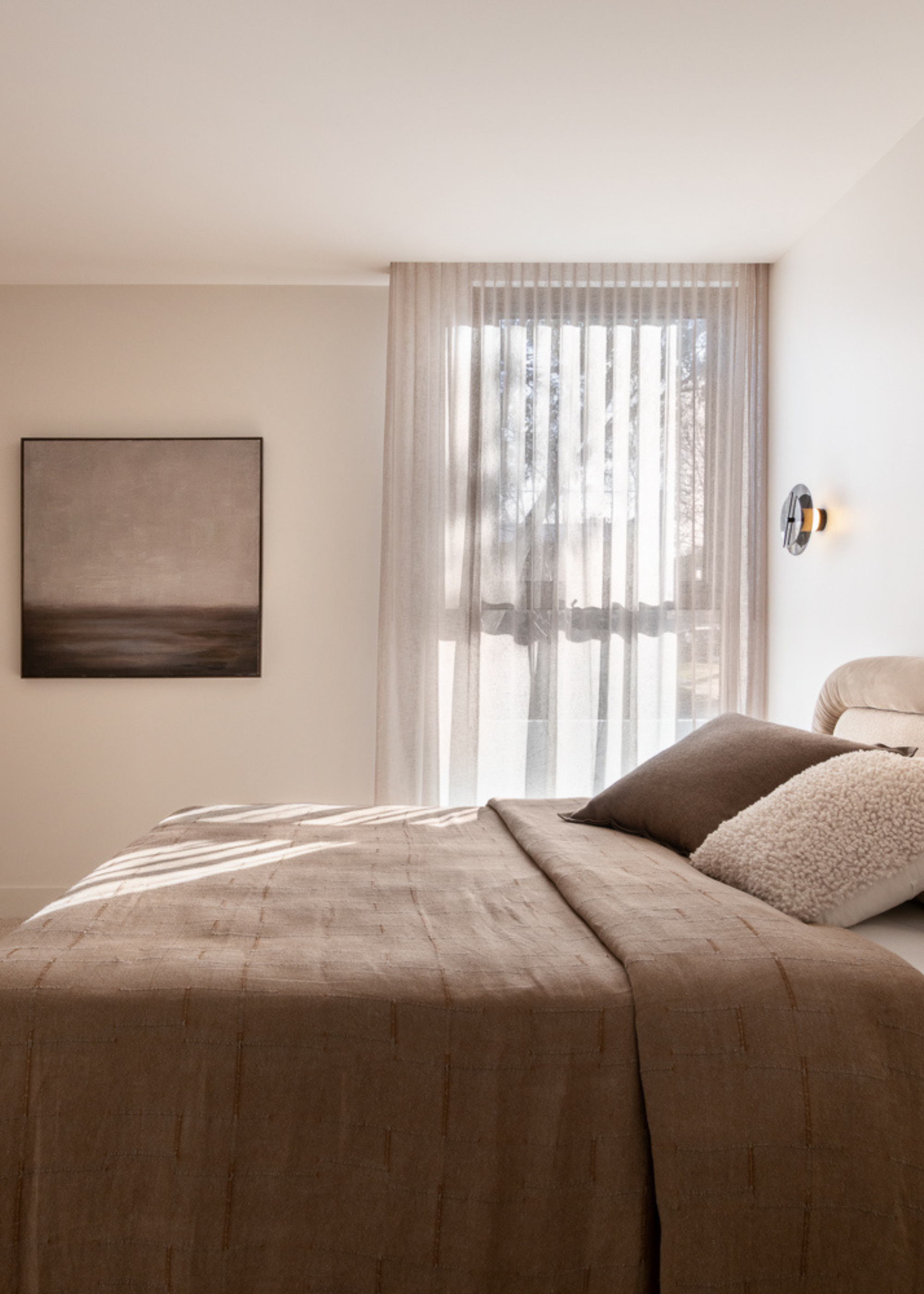 A white bedroom with a framed painting, sheer window treatments, glass wall sconces, and a brown bedscape