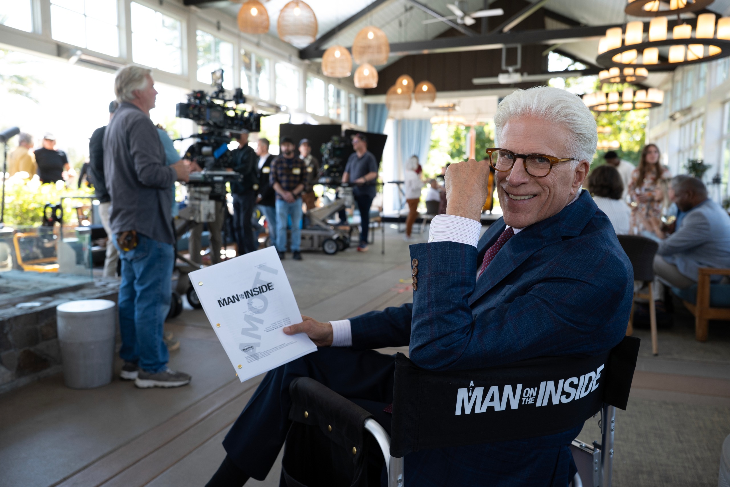 ted danson smiles while holding a script and sitting in a chair on the set of a man on the inside season 3 as the show begins filming
