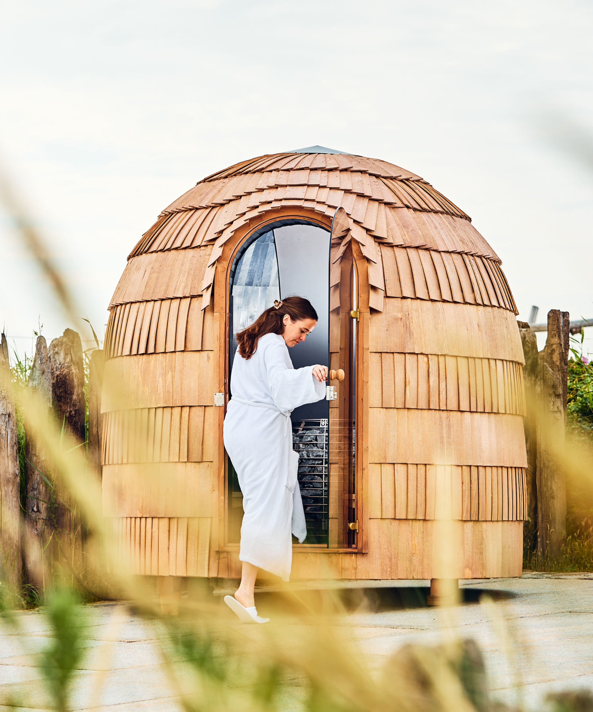 wooden dome style sauna with shingles on exterior, located in garden with female wearing dressing gown stepping inside