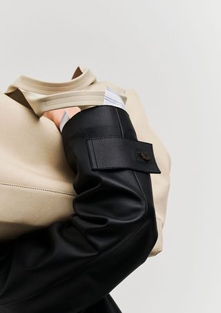 An image of a hand in black leather jacket with a cream tote bag on the shoulder photographed against a grey background