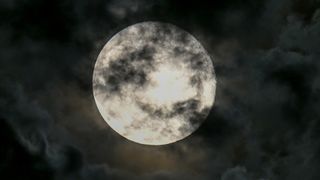 A full moon is shown against a black sky surrounded by clouds, with some encroaching on its surface.