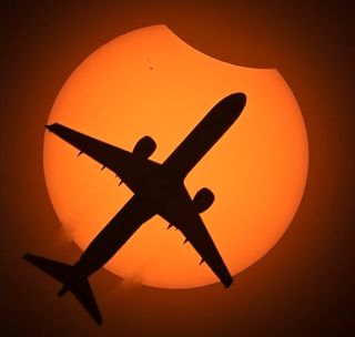 the silhouette of an airplane passes in front of the solar disk which has a chunk missing in the upper right corner where the moon begins to cover it.