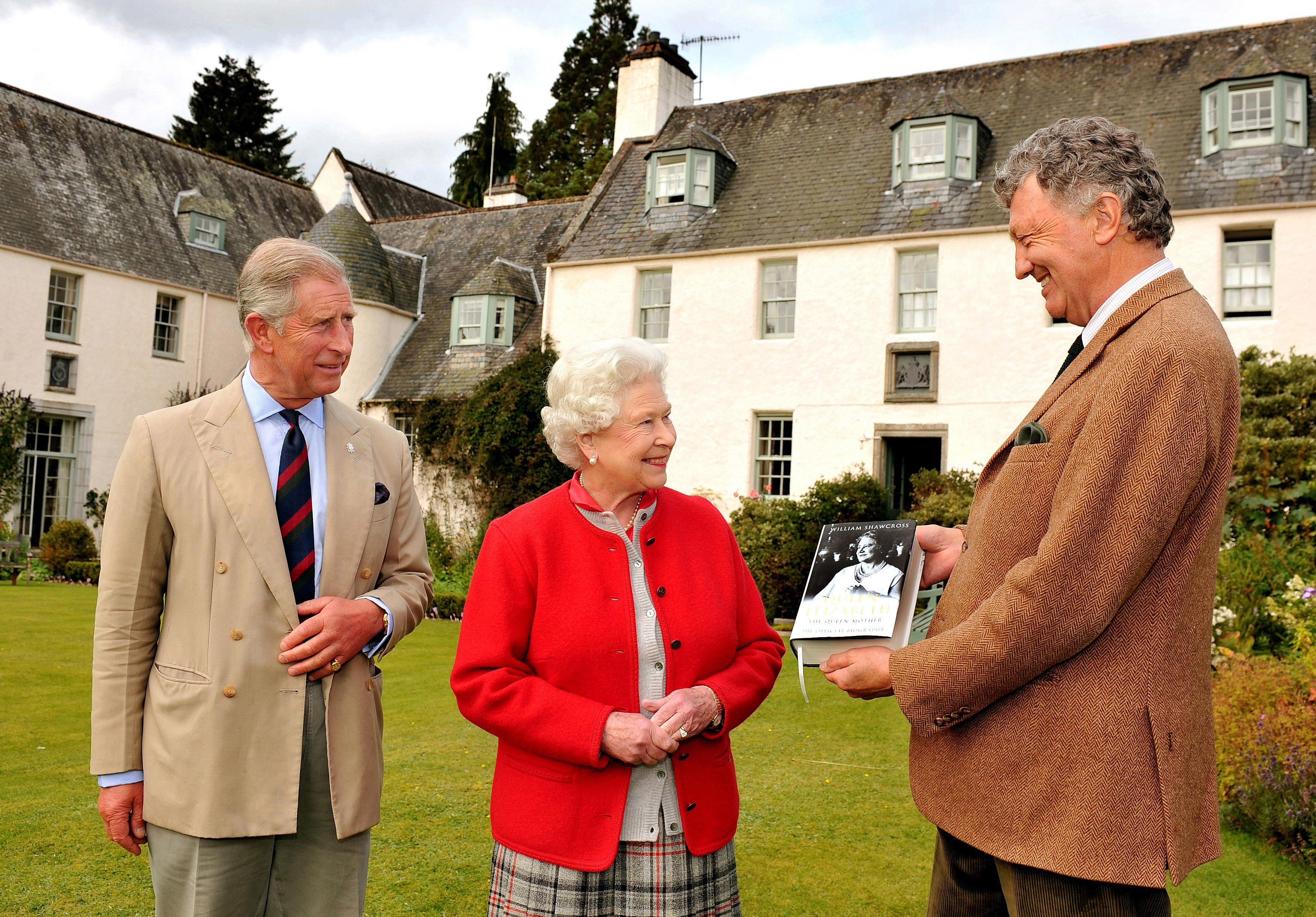 Prince Charles, Queen Elizabeth and William Shawcross stand in the garden of Birkhall as Shawcross presents them with a copy of his biography of the Queen Mother.