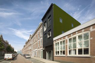 A street with cars parked on both sides. On the right is a terraced building with the Black pearl studio in between. The studio stands out with black bricks and shutter windows in front and the side has windows with green wall grass