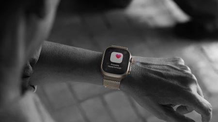 A mostly black-and-white photo of a person wearing the Apple Watch Ultra 3 — with a gold case — with a "Possible Hypertension" pop-up shown in full color.