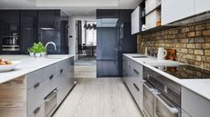 A bright and modern kitchen with white wood floors, grey and blue cabinets, white countertops, and an exposed brick wall.