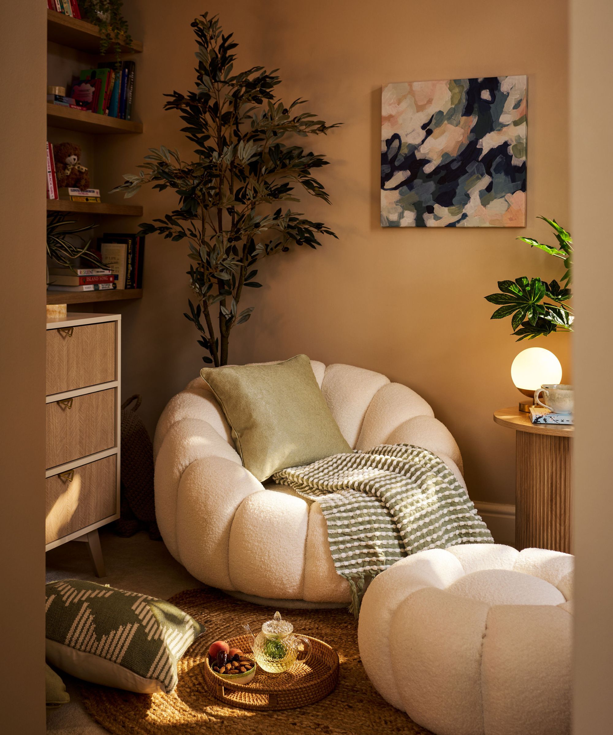 A warm, soothing reading nook featuring a plush cream boucle armchair with matching footstool, soft green accents and a woven rug. Shelving, plants and soft lighting add to the calm, cocooning feel of this intimate corner space.