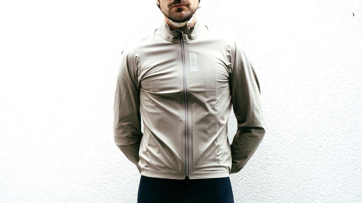 A white man stands against a white wall wearing an off-white waterproof jacket