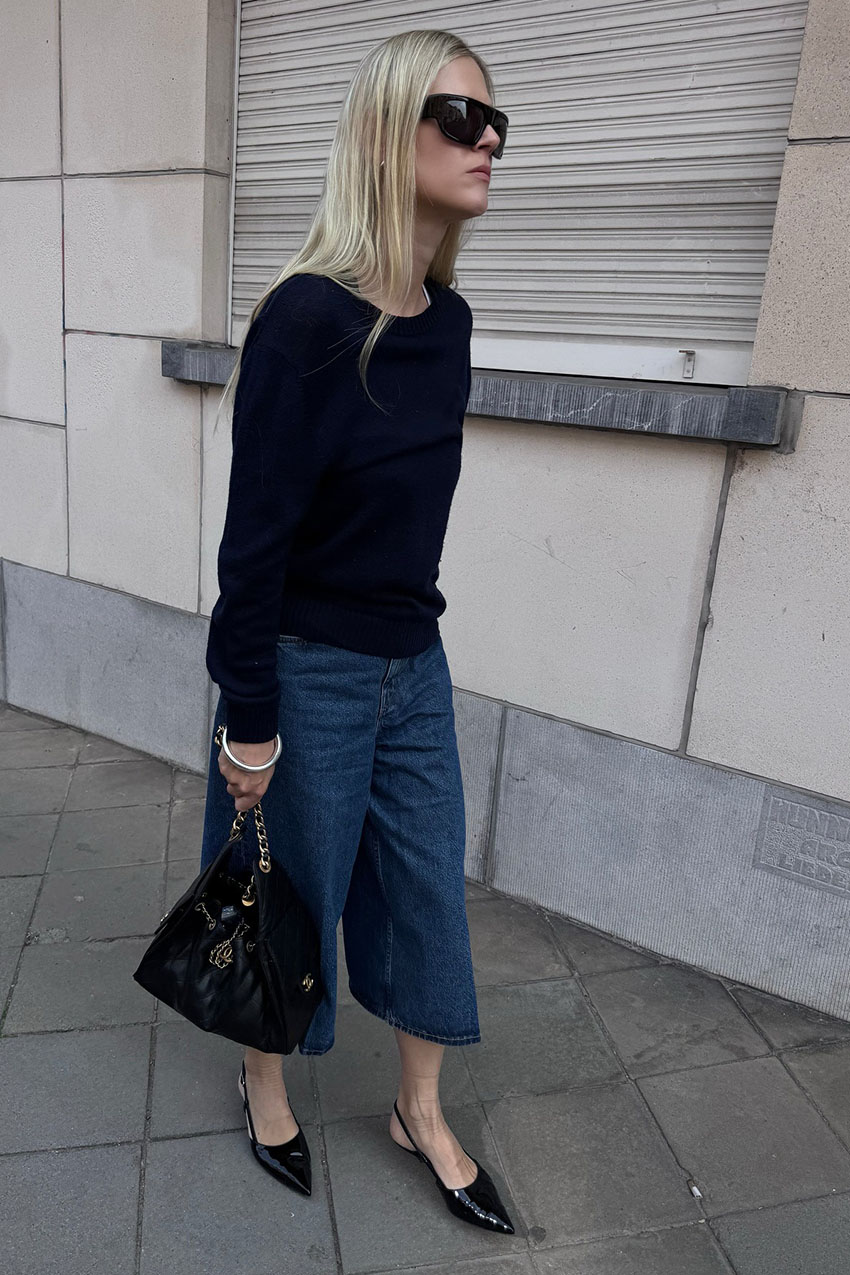 A woman wearing a black sweater, denim culottes, and black pointed-toe heels