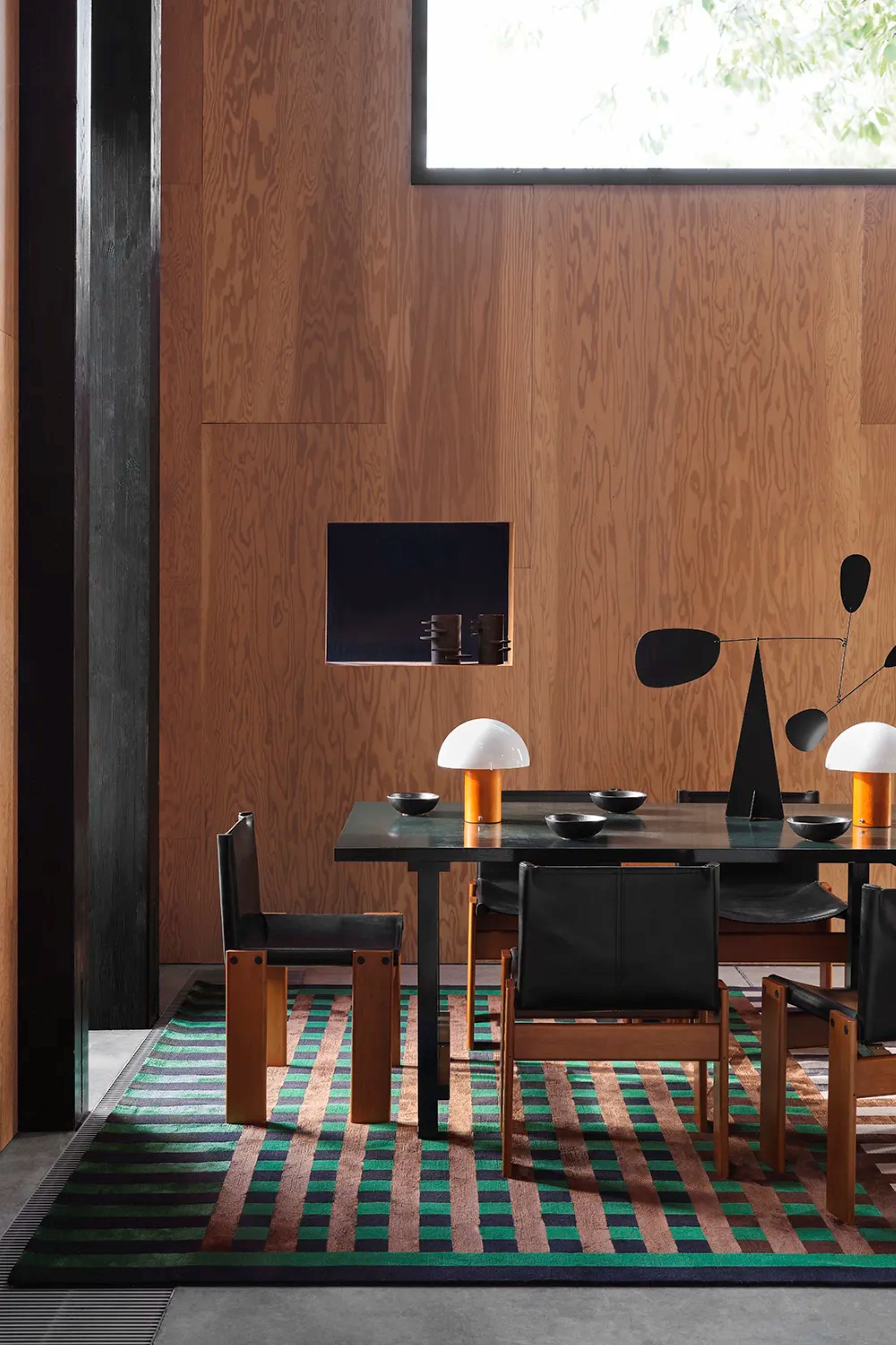 a dining room with a large checker printed rug in green, black, and tan, a large black dining room table, chairs with black leather seats, and a wooden wall