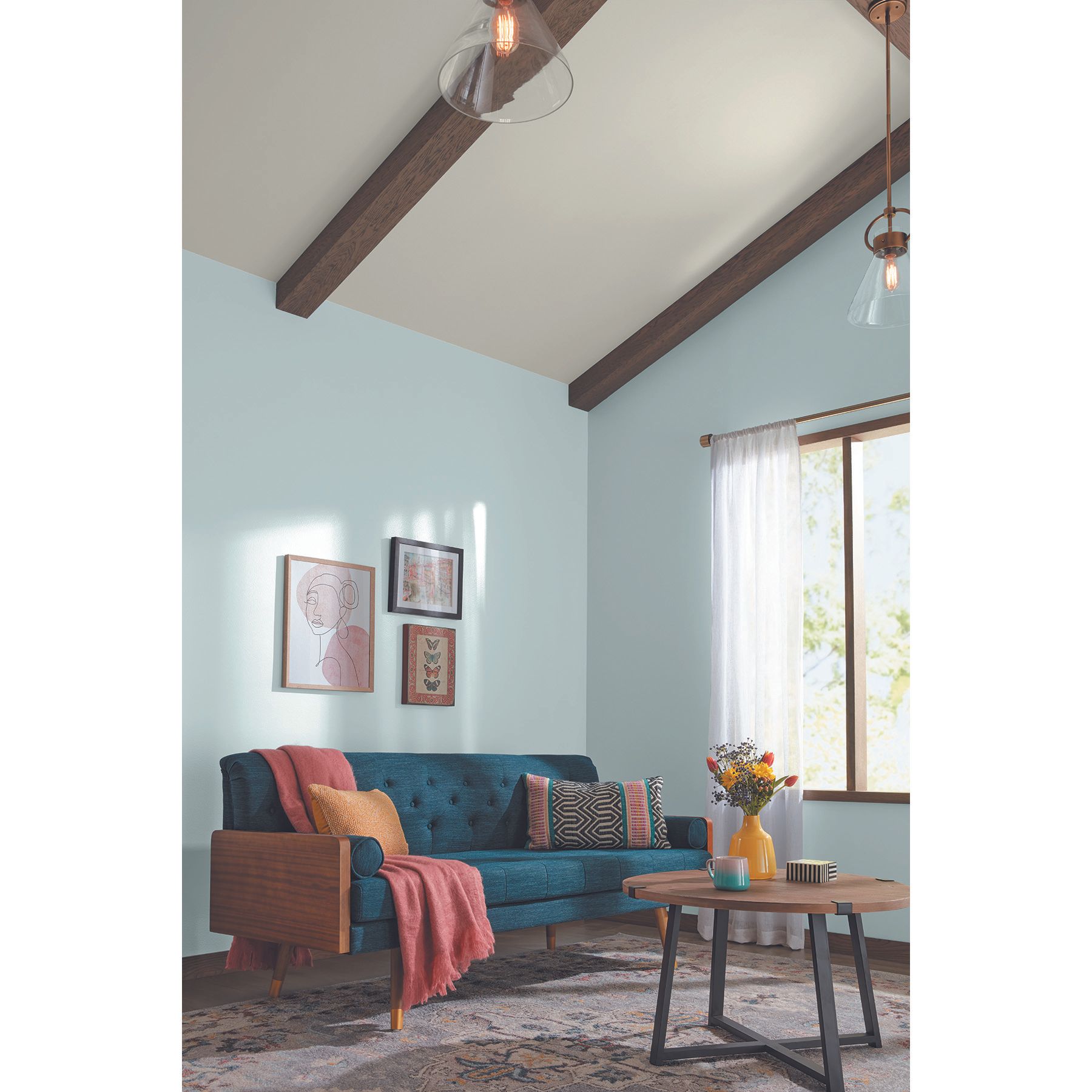 Ruang tamu biru muda dengan sofa biru tua, meja kopi kayu dan logam, permadani antik, karya seni, balok, lemparan karang dan kunyit dan bantal, vas kuning dengan bunga