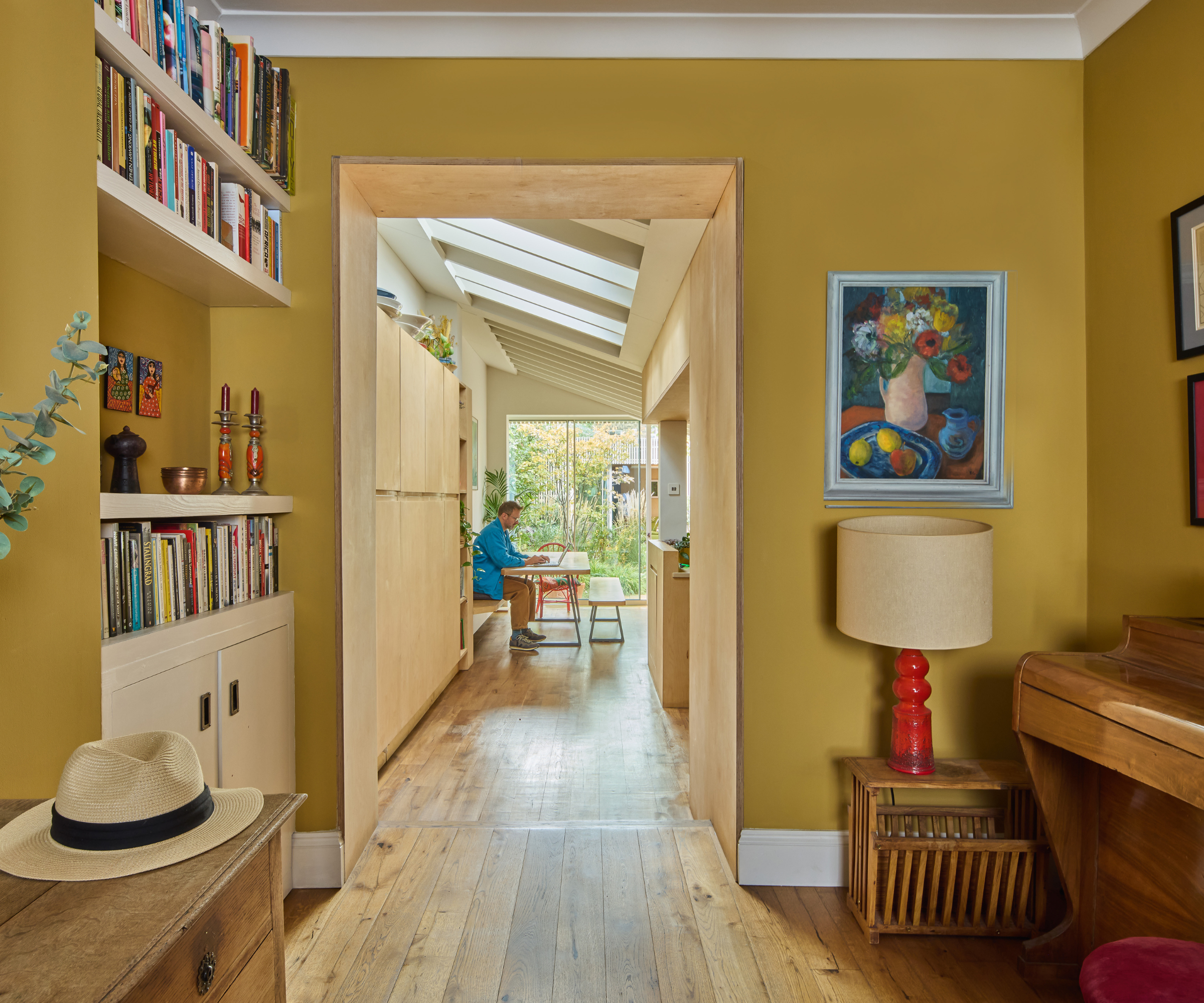 Room with bookshelf to the left and piano and lampshade to the right. Open doorway that looks in to the dining area with a man sitting at the table