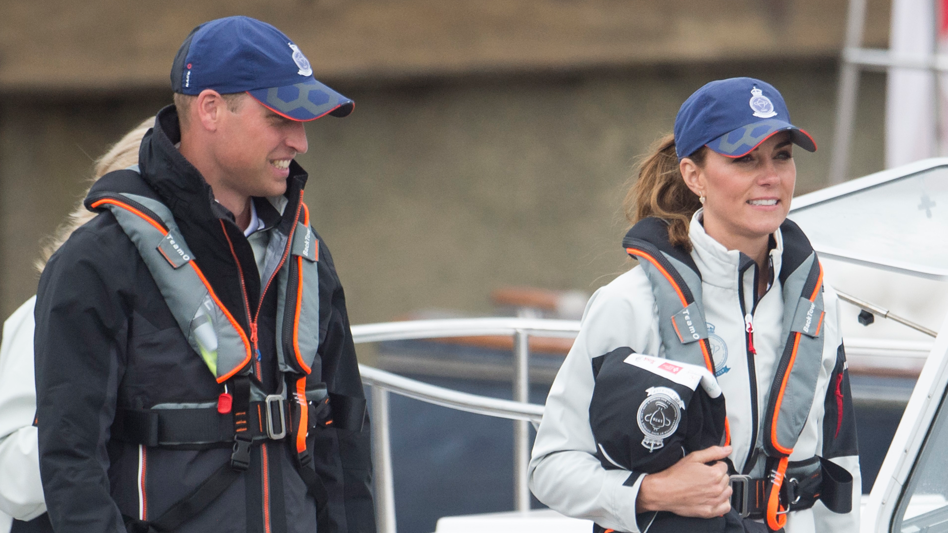 Prince William and Princess Kate wearing blue baseball caps and sailing gear and shorts walking on a dock