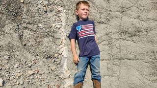 A boy lying next to a partially exposed fossil in the ground.