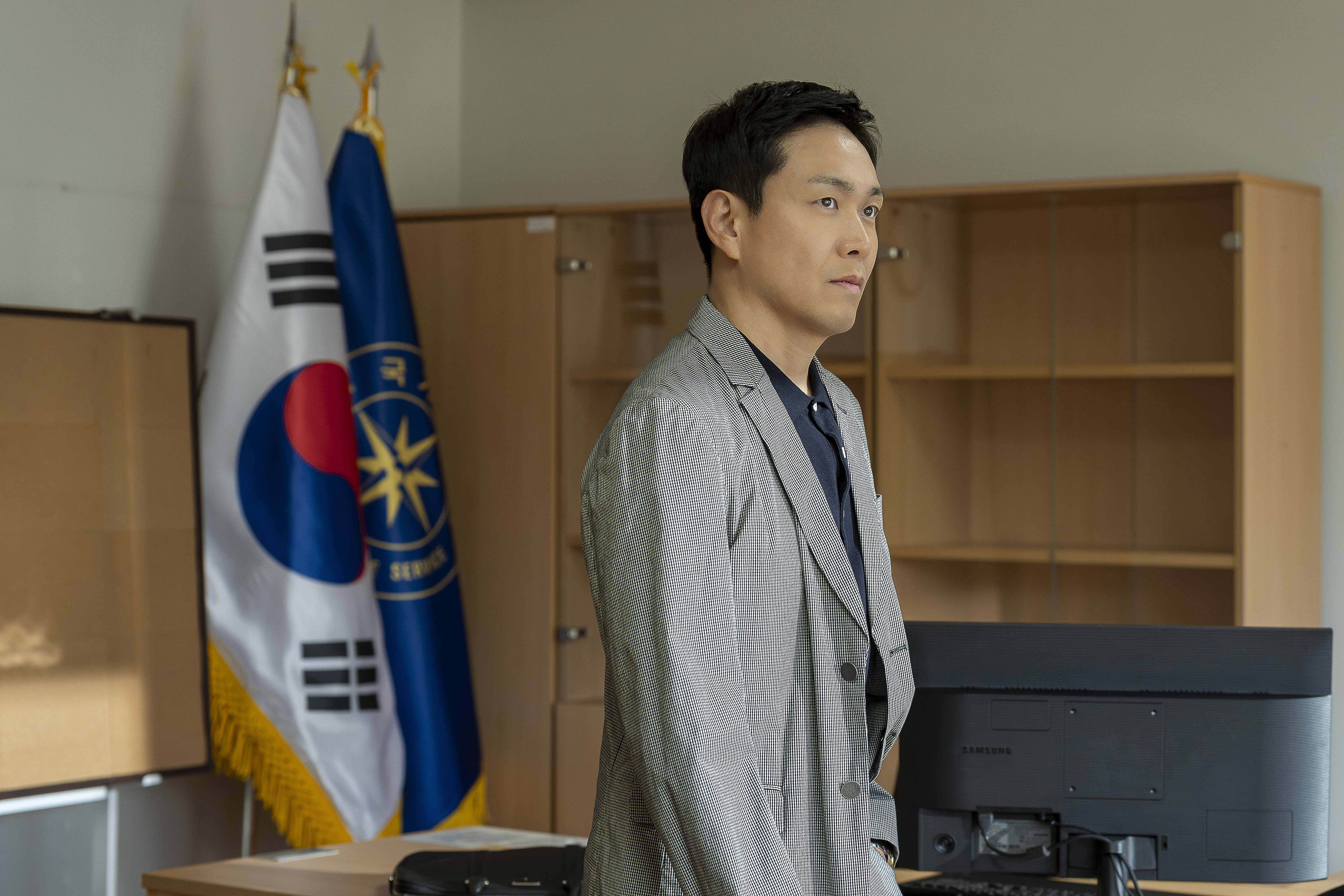Oh Jung-se wears a grey suit and stands in an office with a korean flag in &#039;Tempest.&#039;